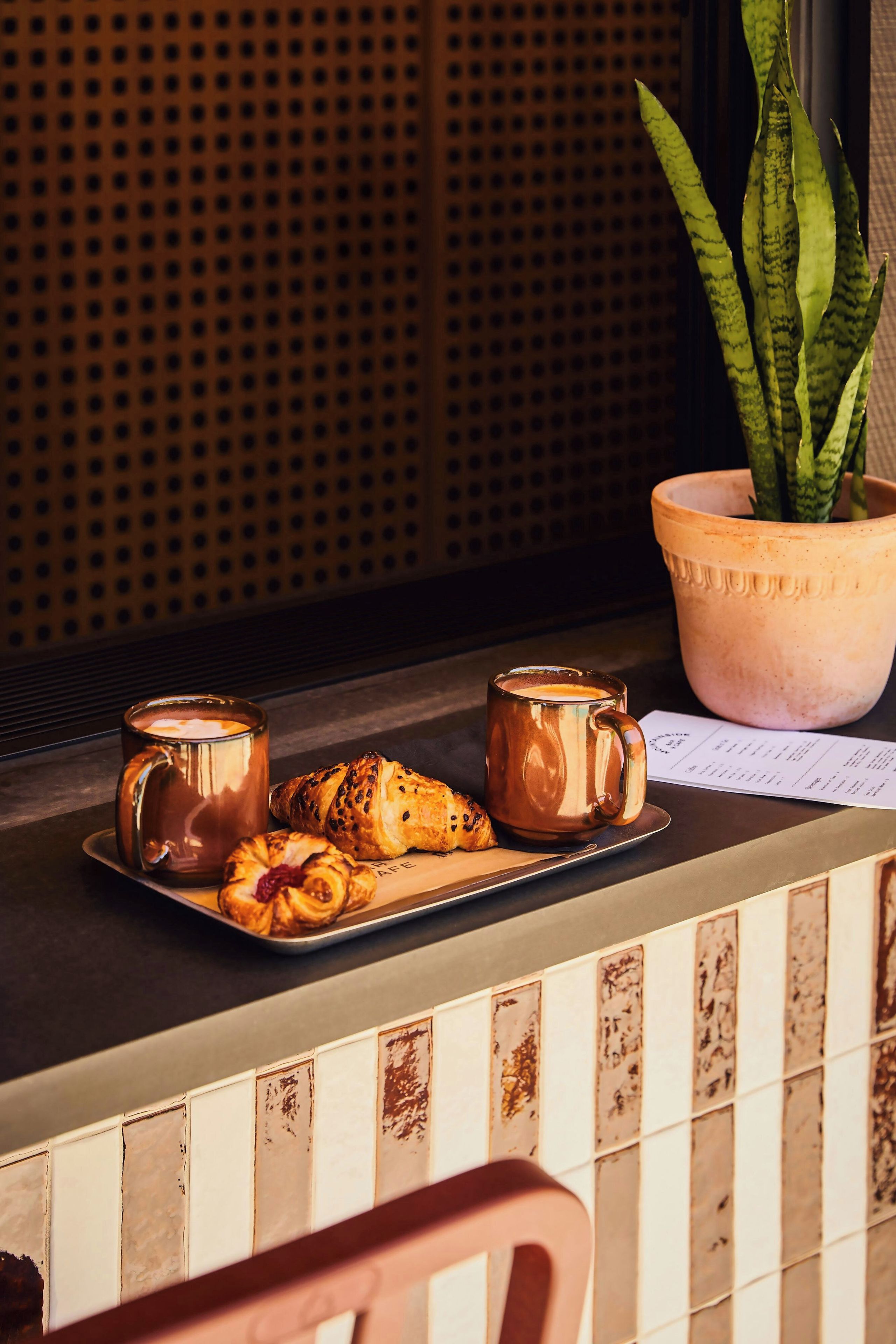 A tray with two mugs and a pastry on a counter, a potted plant, and a tiled wall.