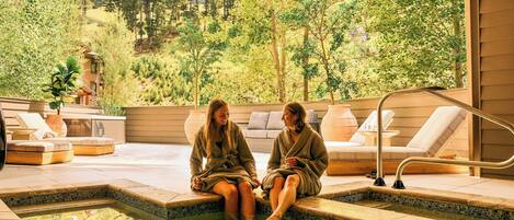 Duas pessoas sentadas à beira de uma piscina, com vista para uma paisagem arborizada.