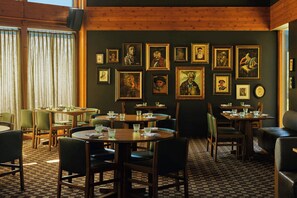 Un restaurant doté de nombreuses tables et chaises, avec des œuvres d’art encadrées accrochées aux murs et un tapis à motifs.
