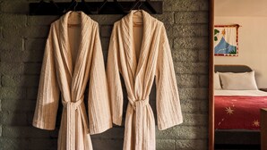 A room with a brick wall, two robes hanging on a rack, a bed with a red blanket, and a painting on the wall.