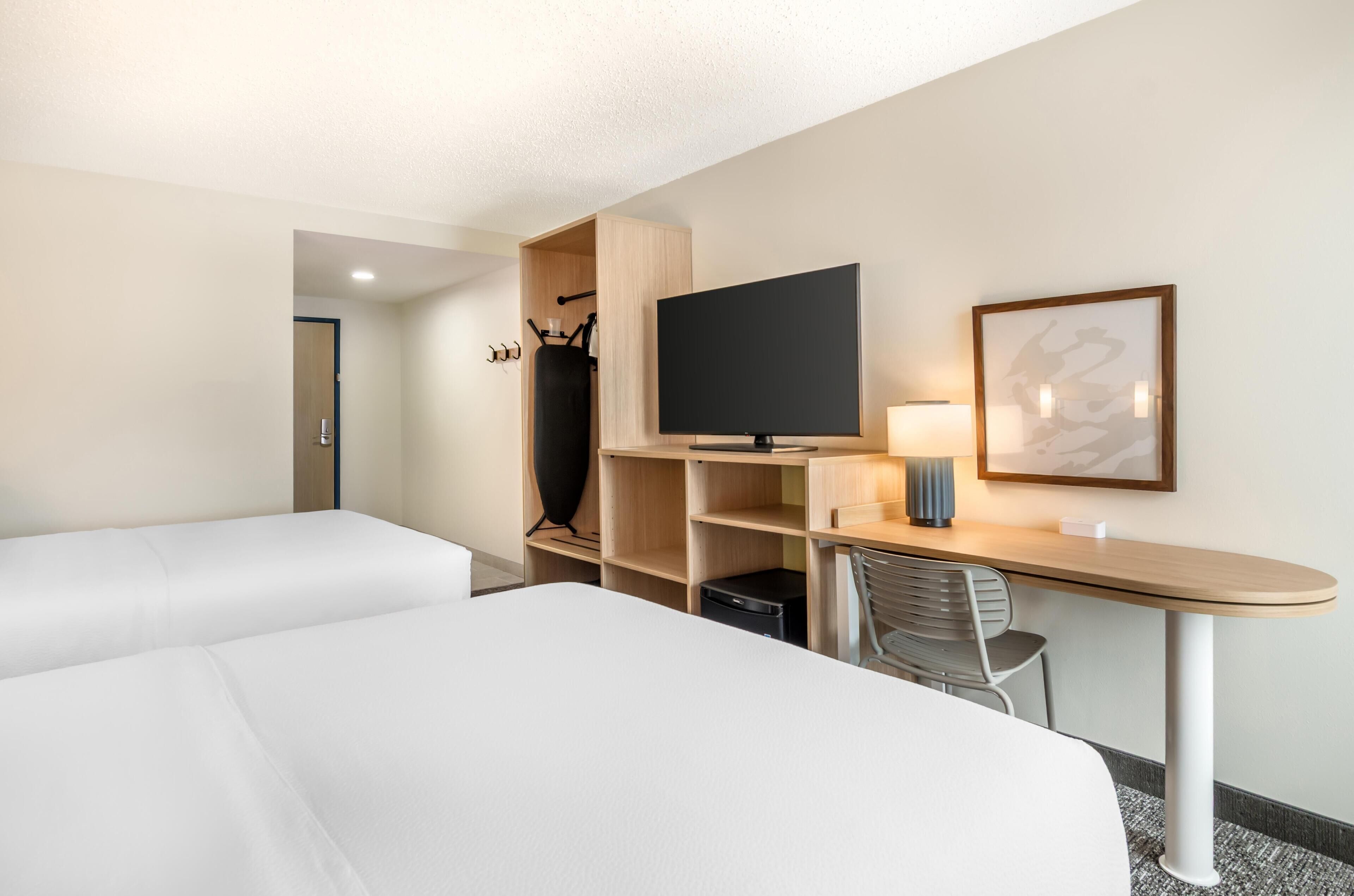 A hotel room with two beds, a TV, a desk with a lamp, and a framed picture on the wall.