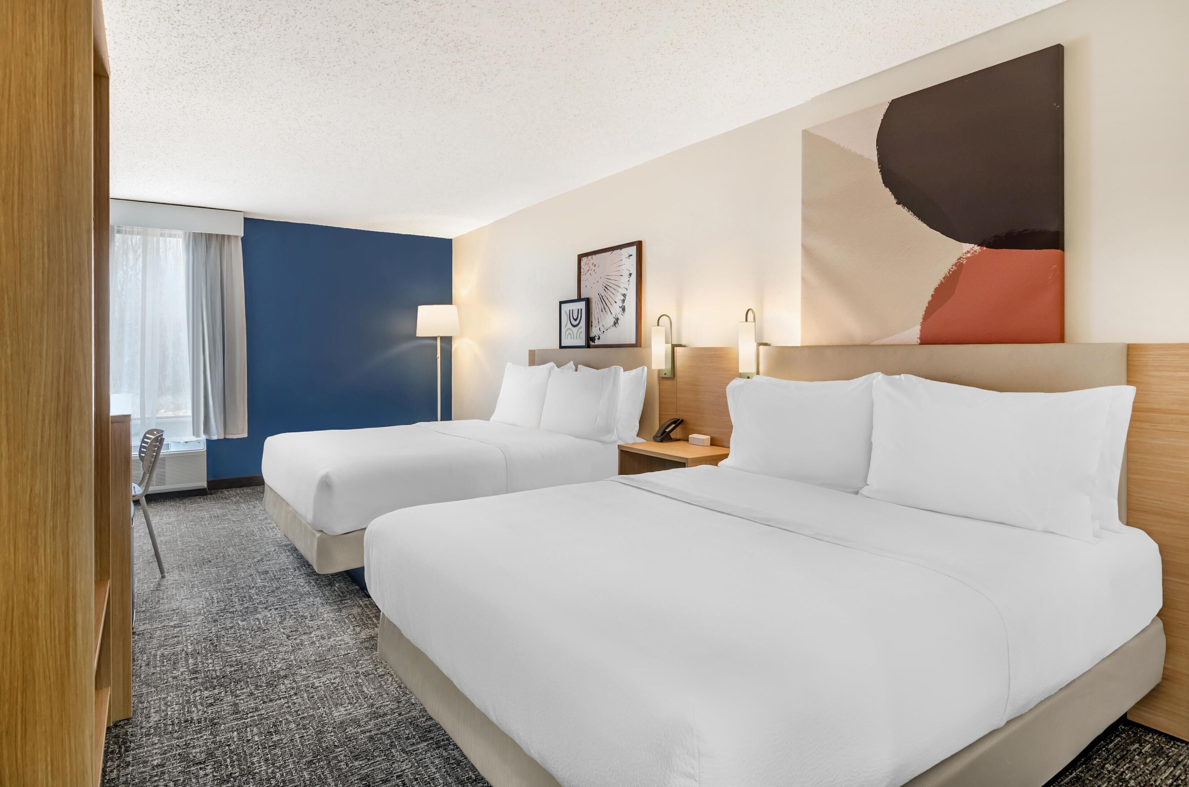 A hotel room with two beds, a large abstract painting, a desk with a lamp, and a window with curtains.