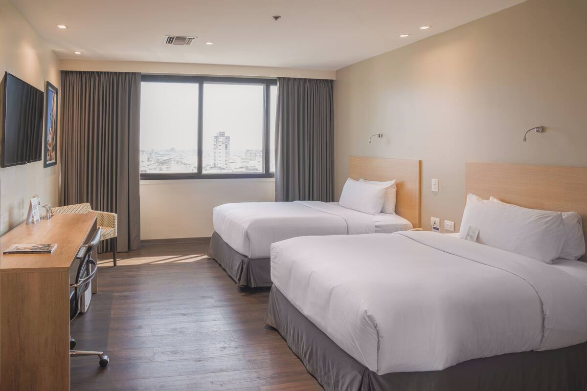 A hotel room with two beds, a desk, and a large window with curtains.
