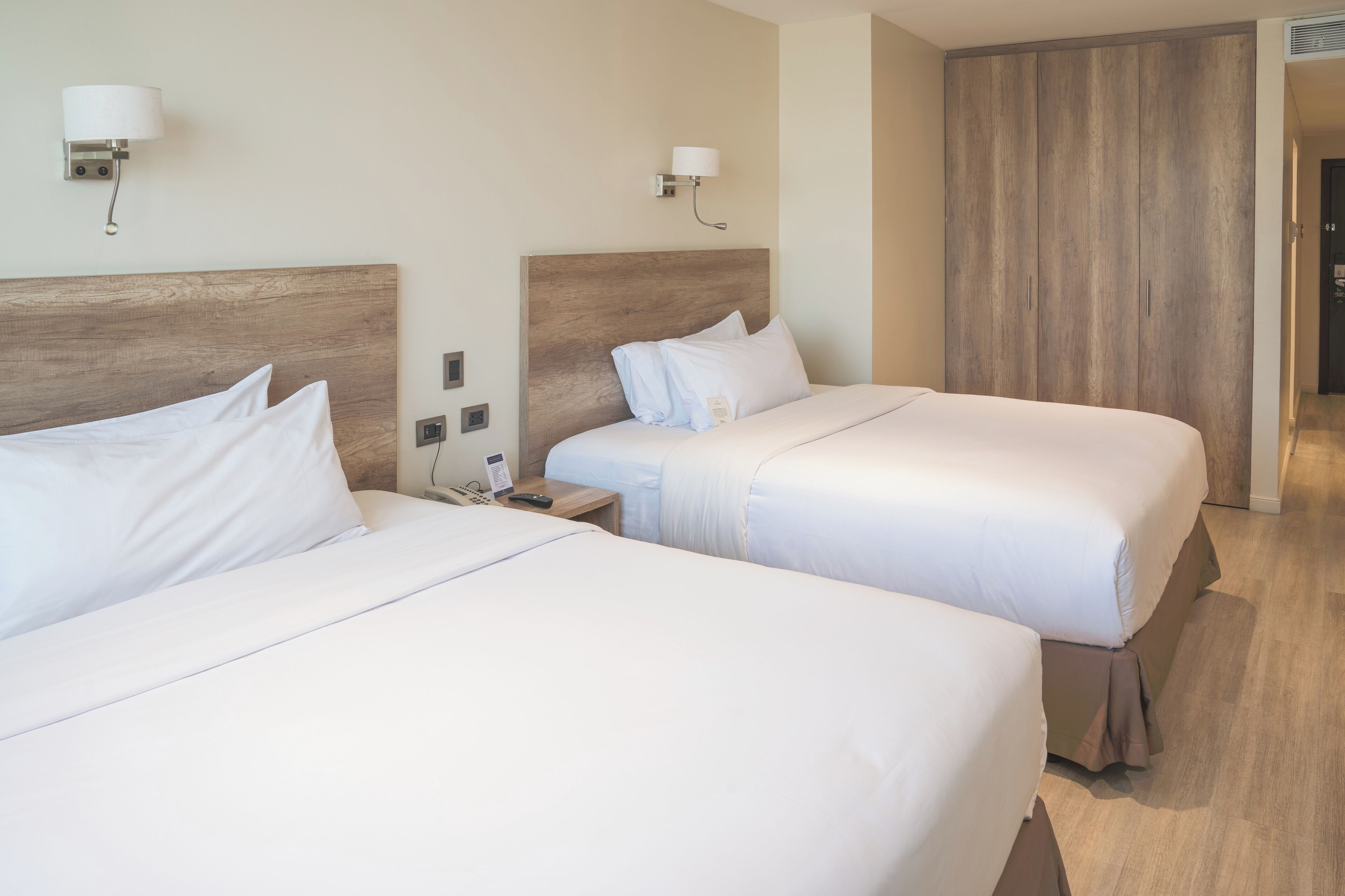 Une chambre d’hôtel avec deux lits, une tête de lit en bois, une table de chevet avec un téléphone et une lampe fixée au mur.
