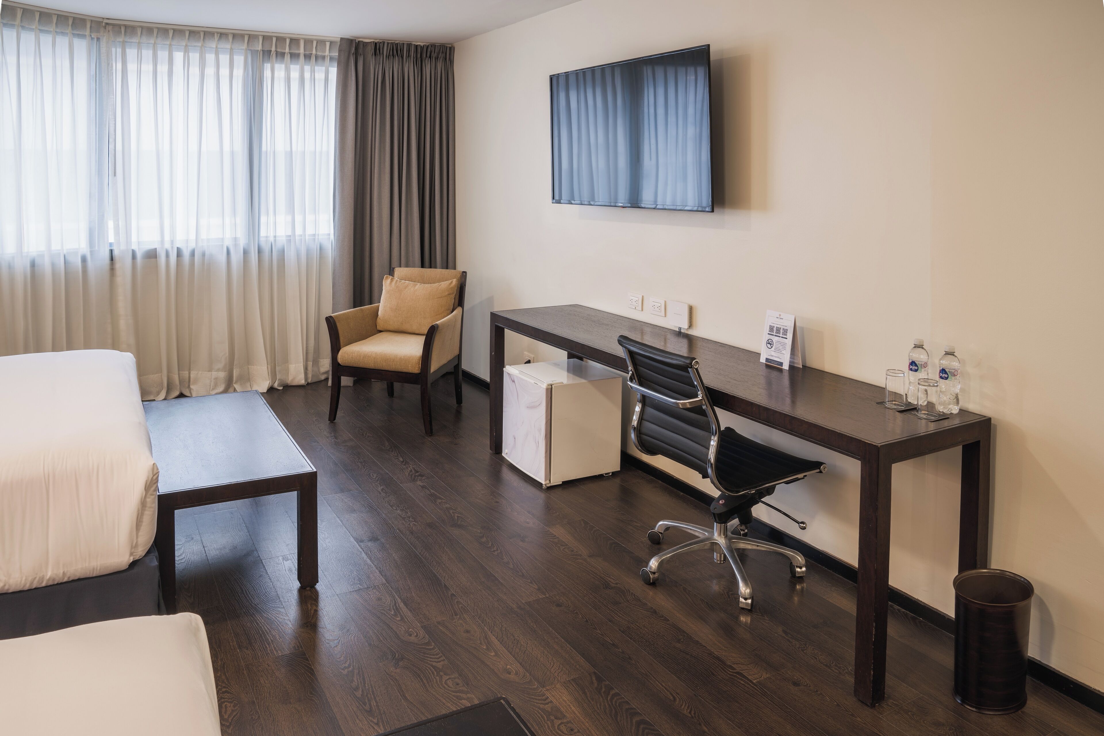 A hotel room with a bed, a desk, a chair, a TV, and a trash can.