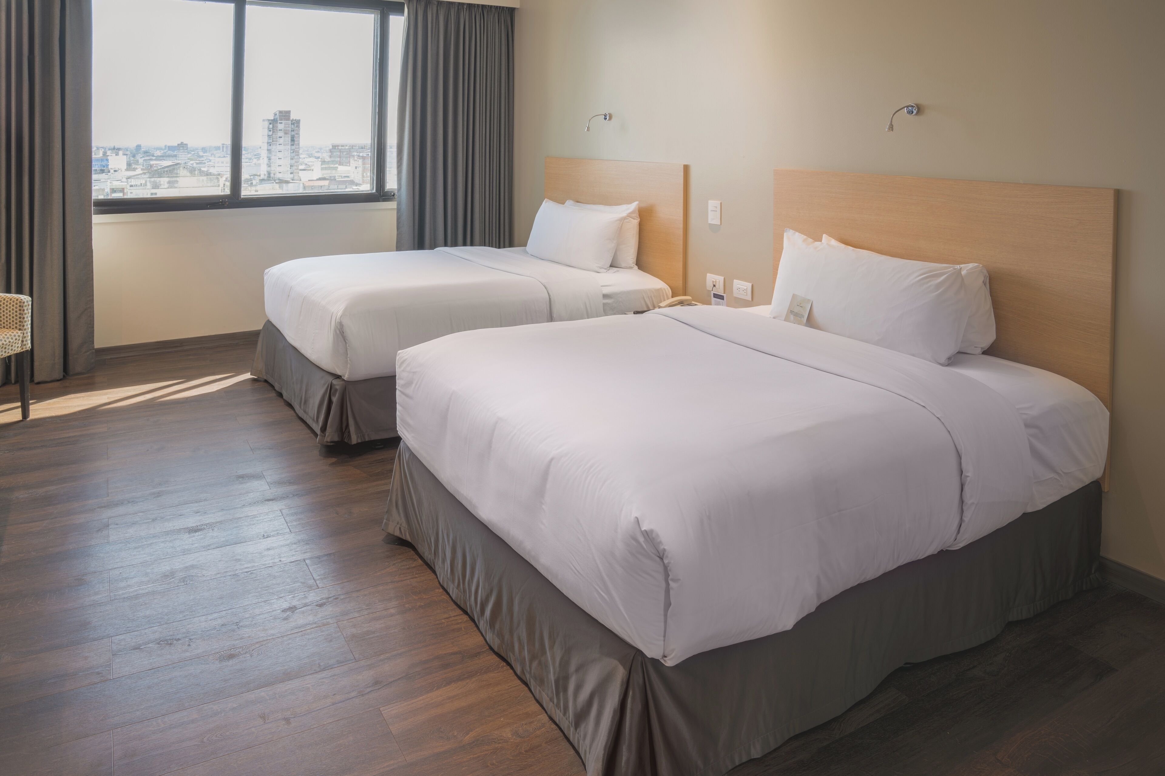 Habitación de hotel con dos camas, un ventanal con cortinas y vista a la ciudad.