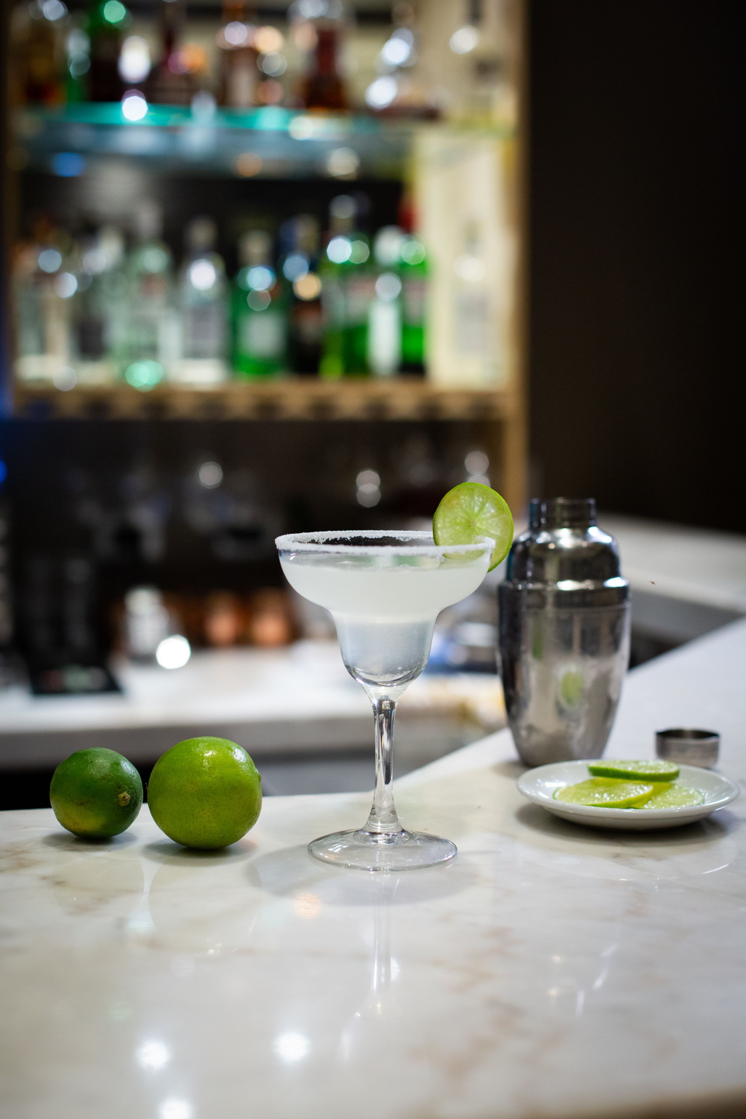 Um copo de martini com um pedaço de limão em um balcão de mármore, dois limões e um coqueteleira ao fundo.