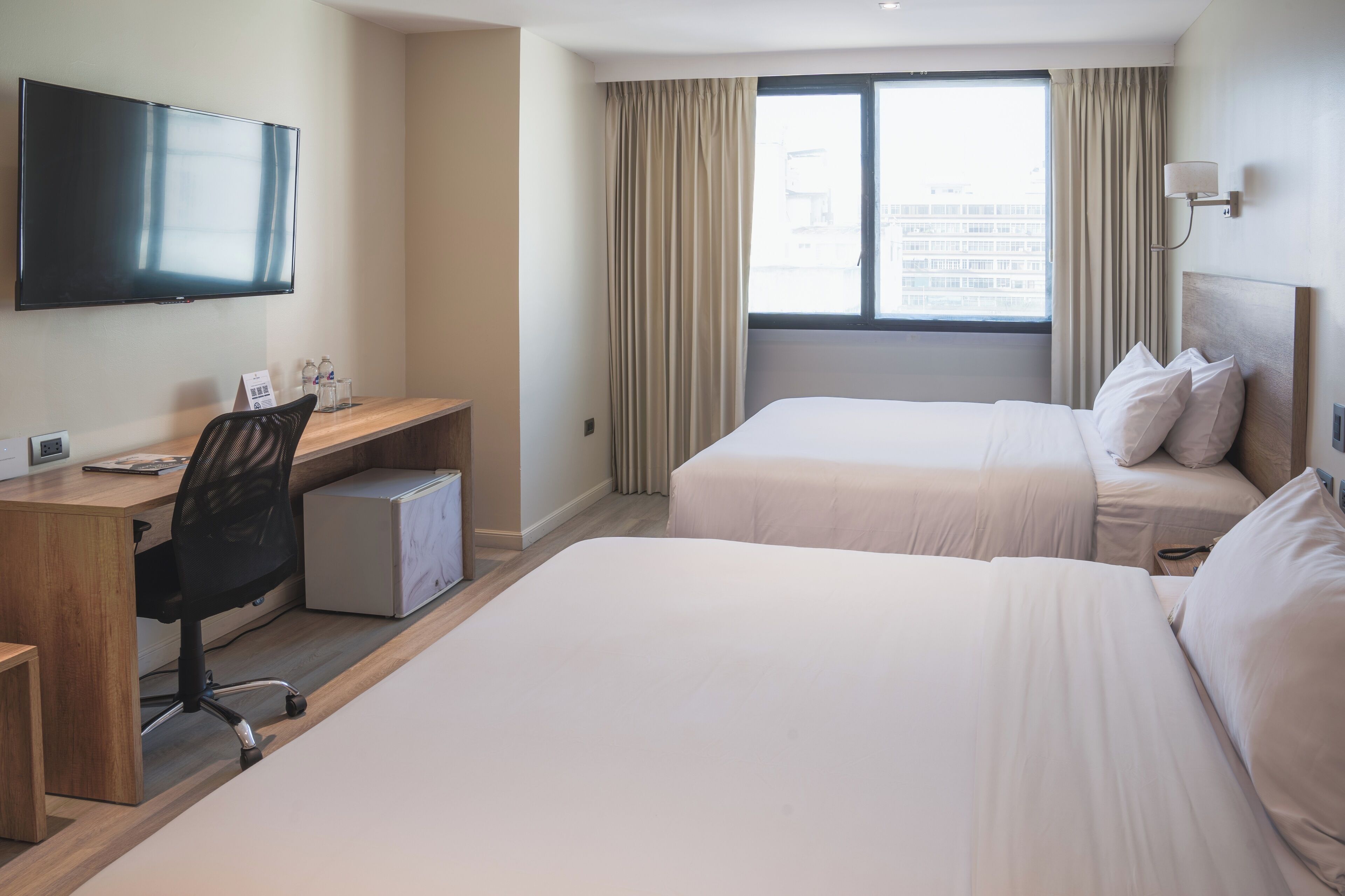 A hotel room with two beds, a desk with a chair, a TV, and a window with curtains.