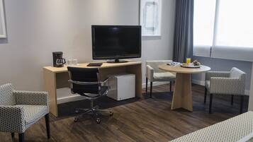 Et hotelværelse med et træskrivebord, et fladskærms-tv, et lille køleskab, to mønstrede stole, et rundt bord med en tallerken med frugt og et glas appelsinjuice, samt en seng med et mønstret sengetæppe.