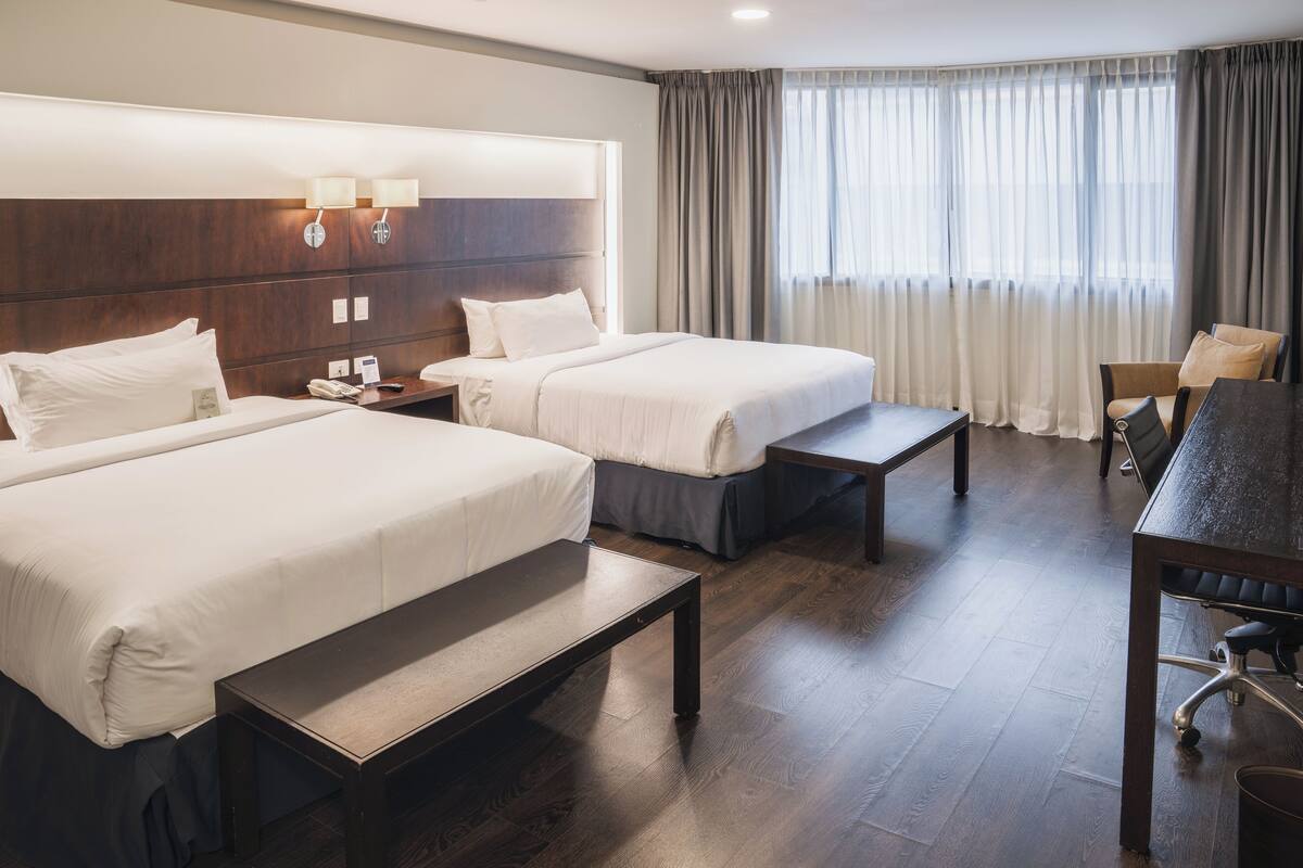 A hotel room with two beds, a desk, a chair, a nightstand, and a lamp.