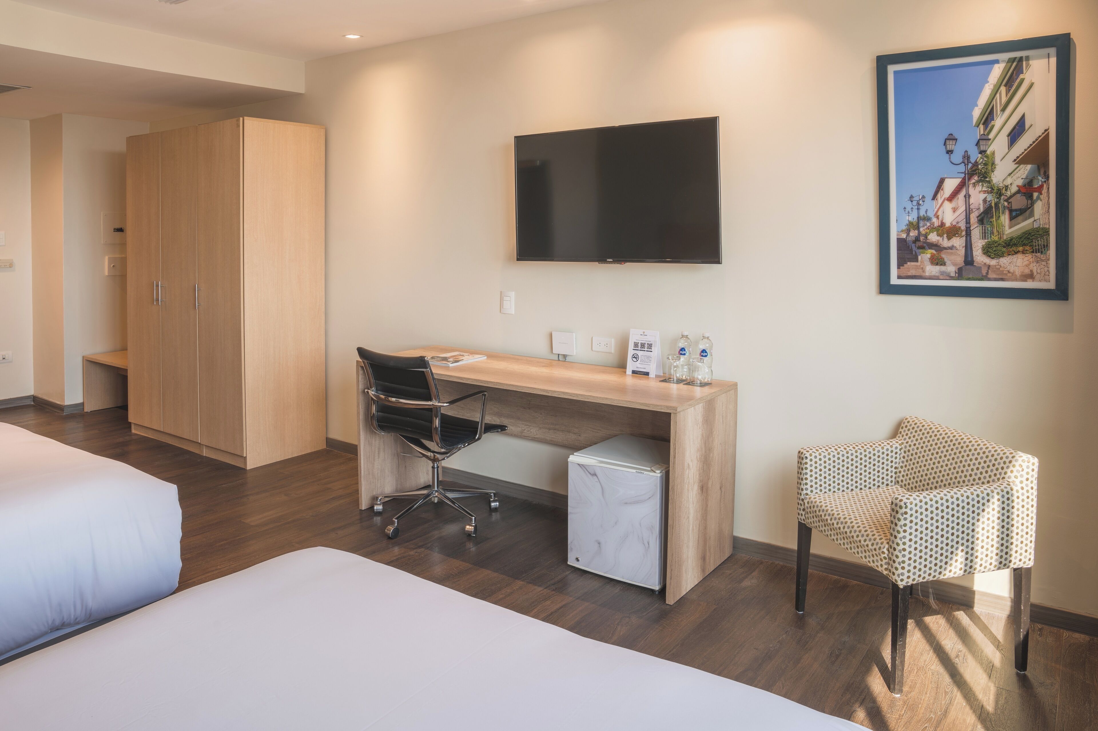 A hotel room with a wooden desk, a chair, a bed, a wardrobe, a television, and a framed picture on the wall.