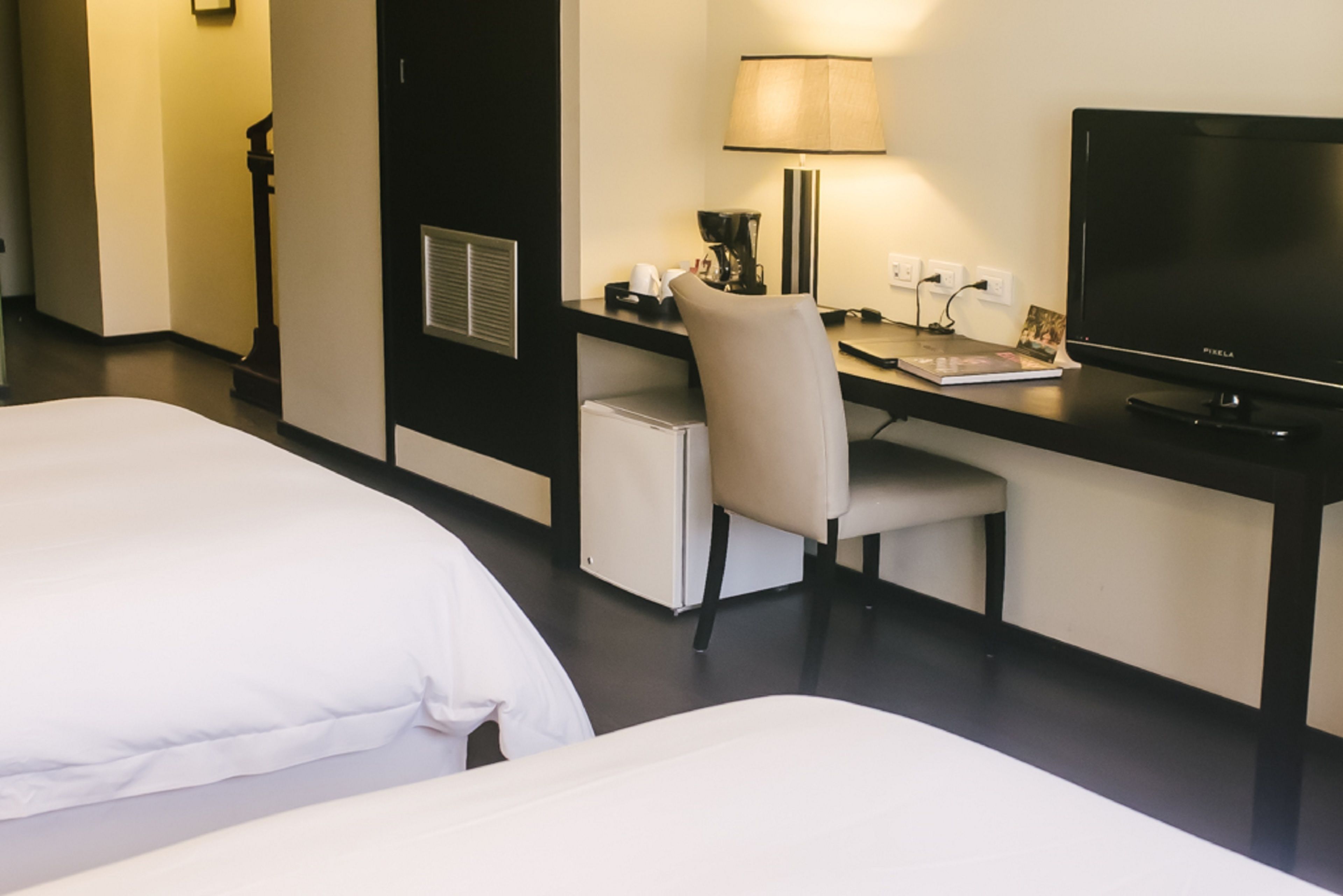 Quarto de hotel com duas camas, uma mesa de trabalho com cadeira, luminária, televisão e cafeteira.