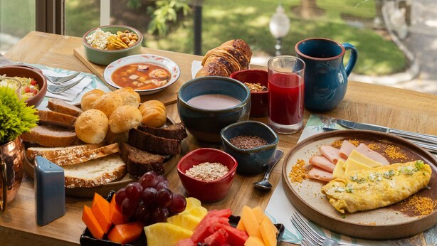 A breakfast table with various foods and a view of a park through a window. - Unipark by Oro Verde Hotels (Guayaquil)