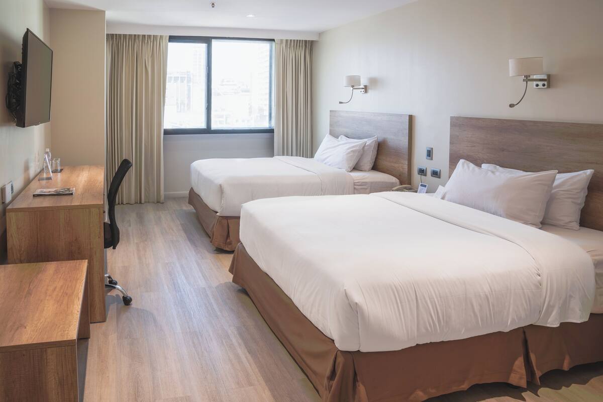 A hotel room with two beds, a desk, a chair, a TV, and a window with curtains.