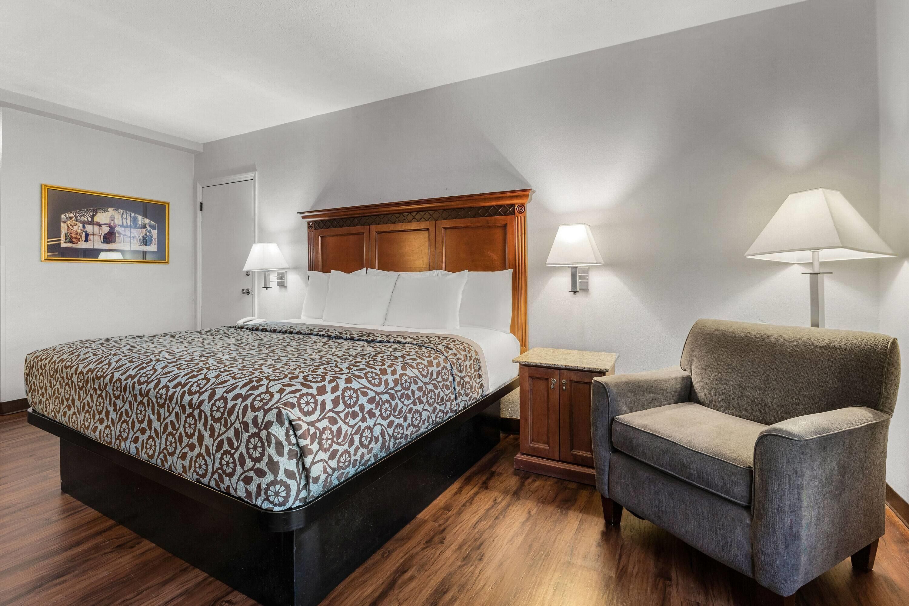 Habitación de hotel con una cama grande, dos lámparas de noche, un cabecero de madera, un sillón gris y una mesita pequeña.