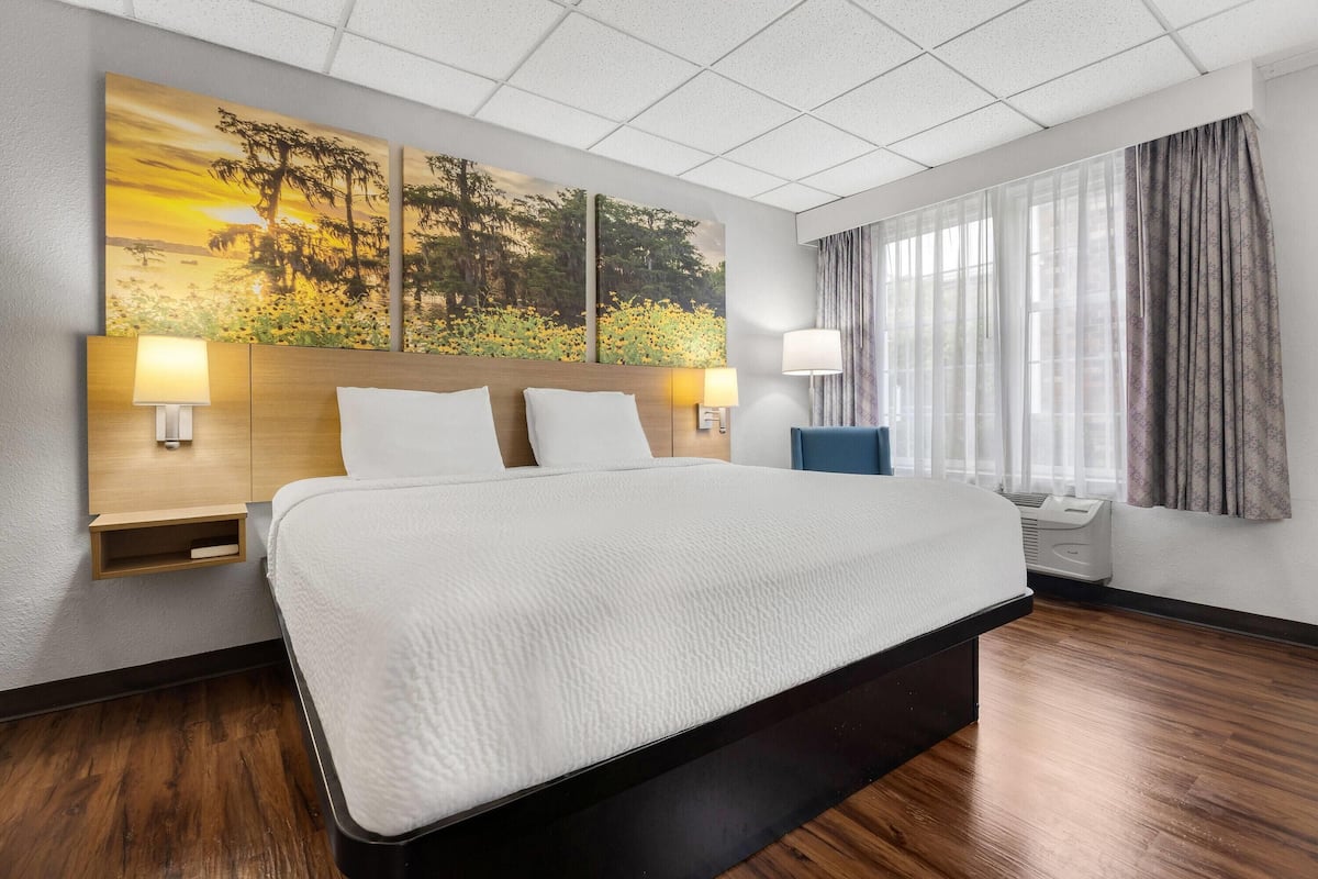 A hotel room with a large bed, two bedside tables with lamps, a window with curtains, and a wall-mounted air conditioning unit.