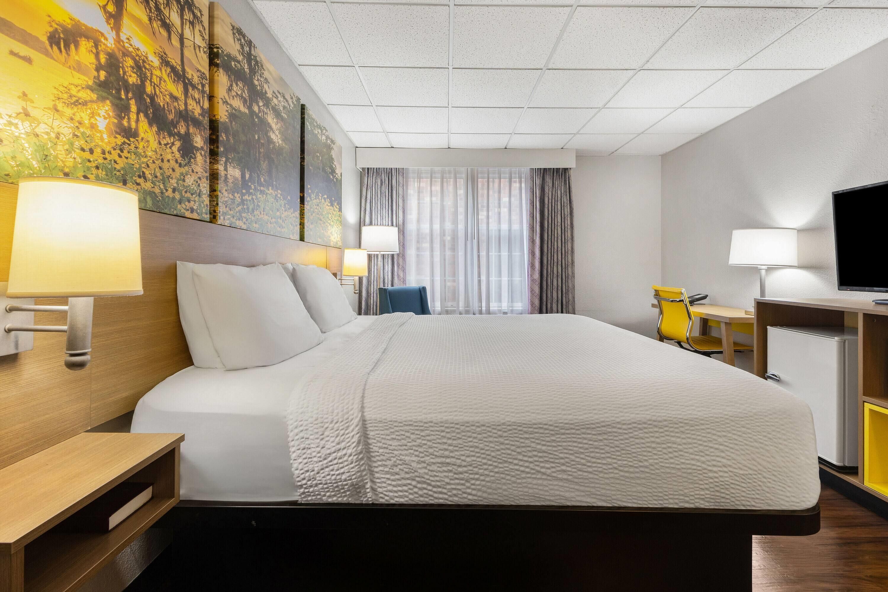 A hotel room with a large bed, two bedside lamps, a desk with a chair, a television, and a window with curtains.