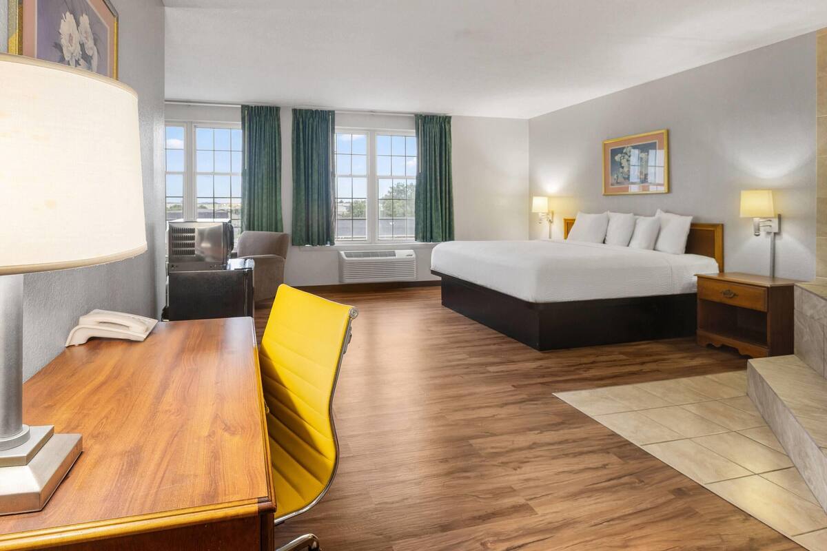 A hotel room with a bed, bedside tables, a desk, a chair, a lamp, and a window with curtains.