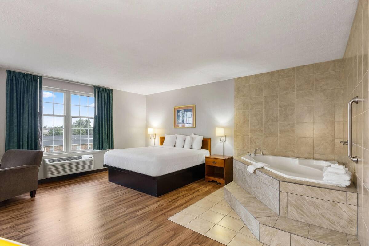 A hotel room with a large bed, a sitting area, a window with curtains, a wall-mounted picture, a bathroom with a bathtub, and a handrail.