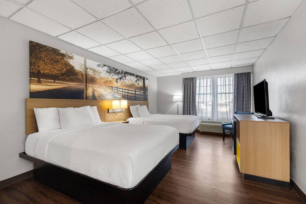 A hotel room with two beds, a desk, a TV, and a large window with curtains.