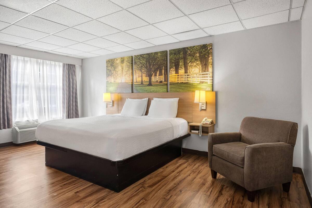 A hotel room with a large bed, a chair, a painting on the wall, and a window with curtains.