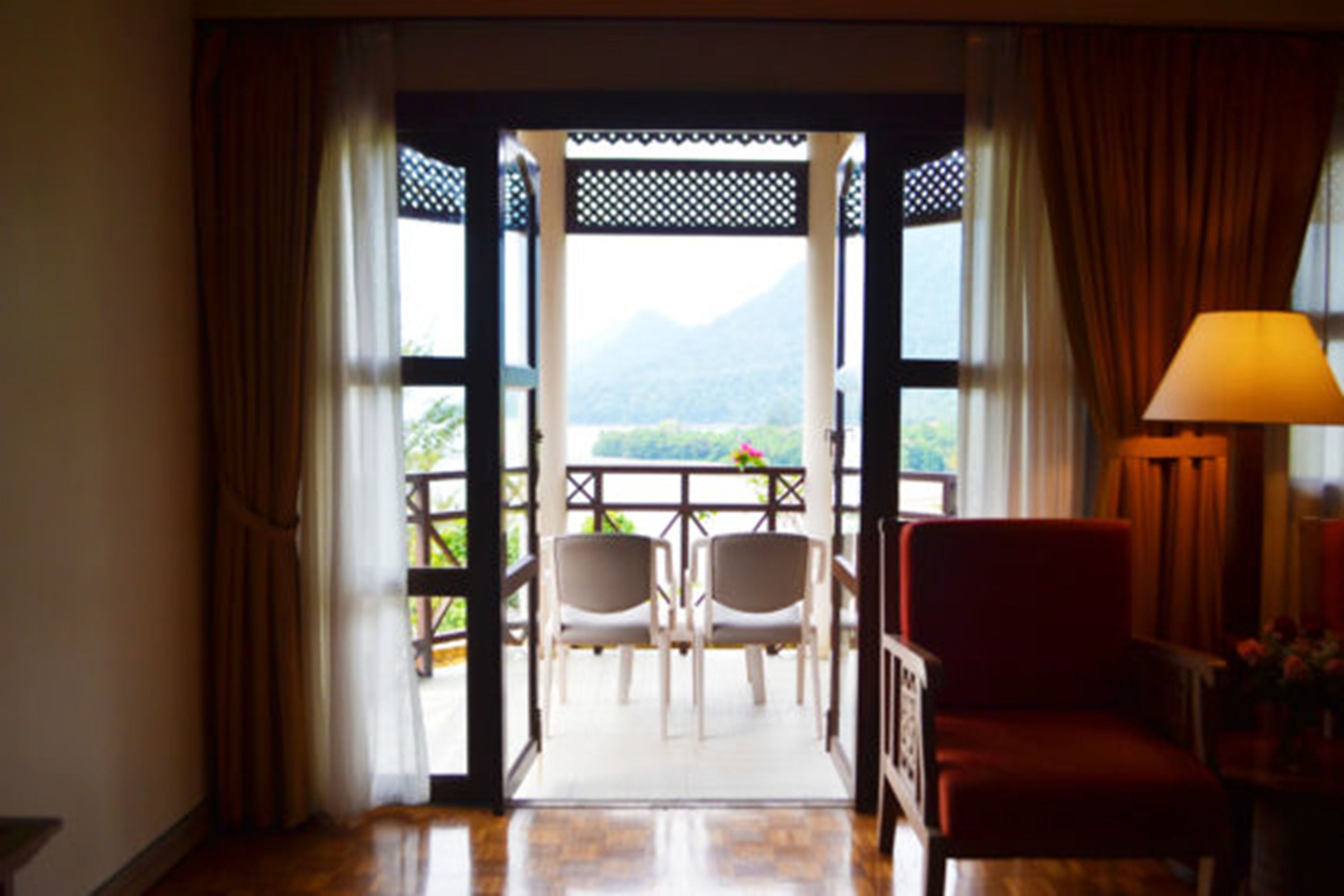 An open doorway leading to a balcony with two chairs and a table, a red chair inside, and a lamp on a side table.