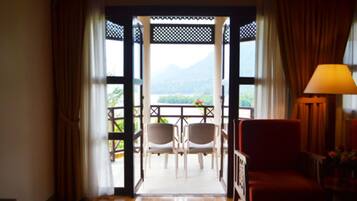 An open doorway leading to a balcony with two chairs and a table, a red chair inside, and a lamp on a side table.