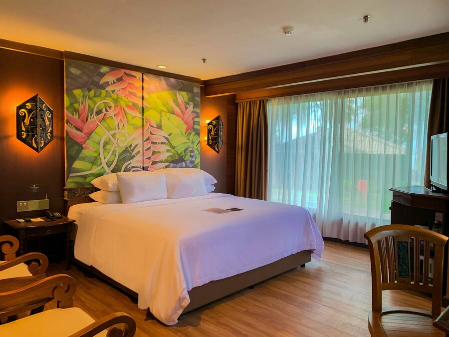 Une chambre d’hôtel avec un grand lit, un bureau, une chaise, un tableau au mur et une fenêtre avec des rideaux.
