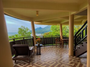 A covered balcony with a tiled floor, wooden furniture, and a view of a mountainous landscape and water.