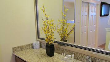 Un baño con encimera de granito, un lavabo blanco, un espejo, un florero con flores amarillas y un televisor montado en la pared.