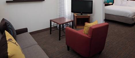A hotel room with a bed, a TV, a small table, a sofa, and a wall-mounted shelf with framed pictures.