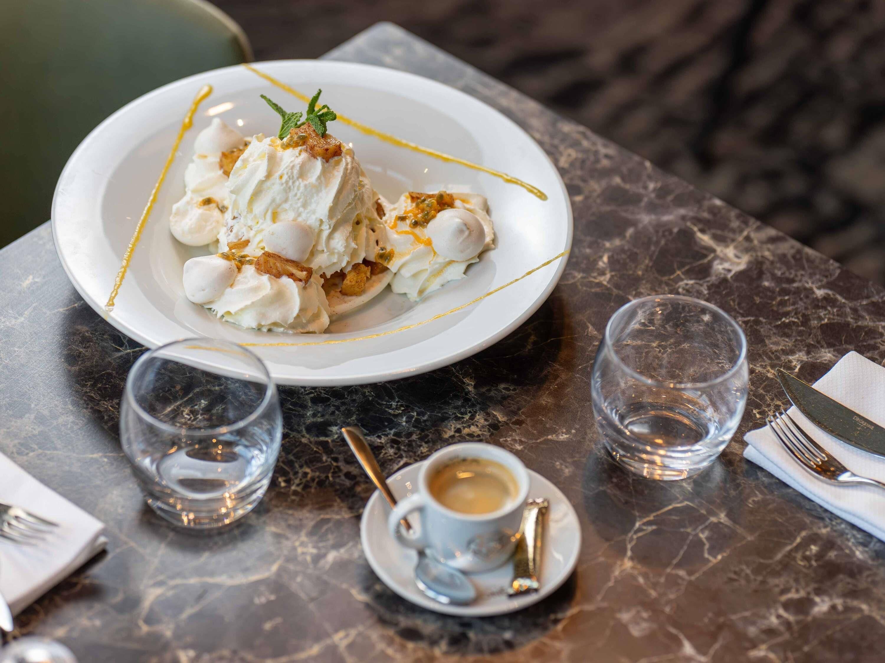 En desserttallrik med vispad grädde och karamelliserad frukt står på ett marmorbord, tillsammans med en kopp kaffe och två glas.