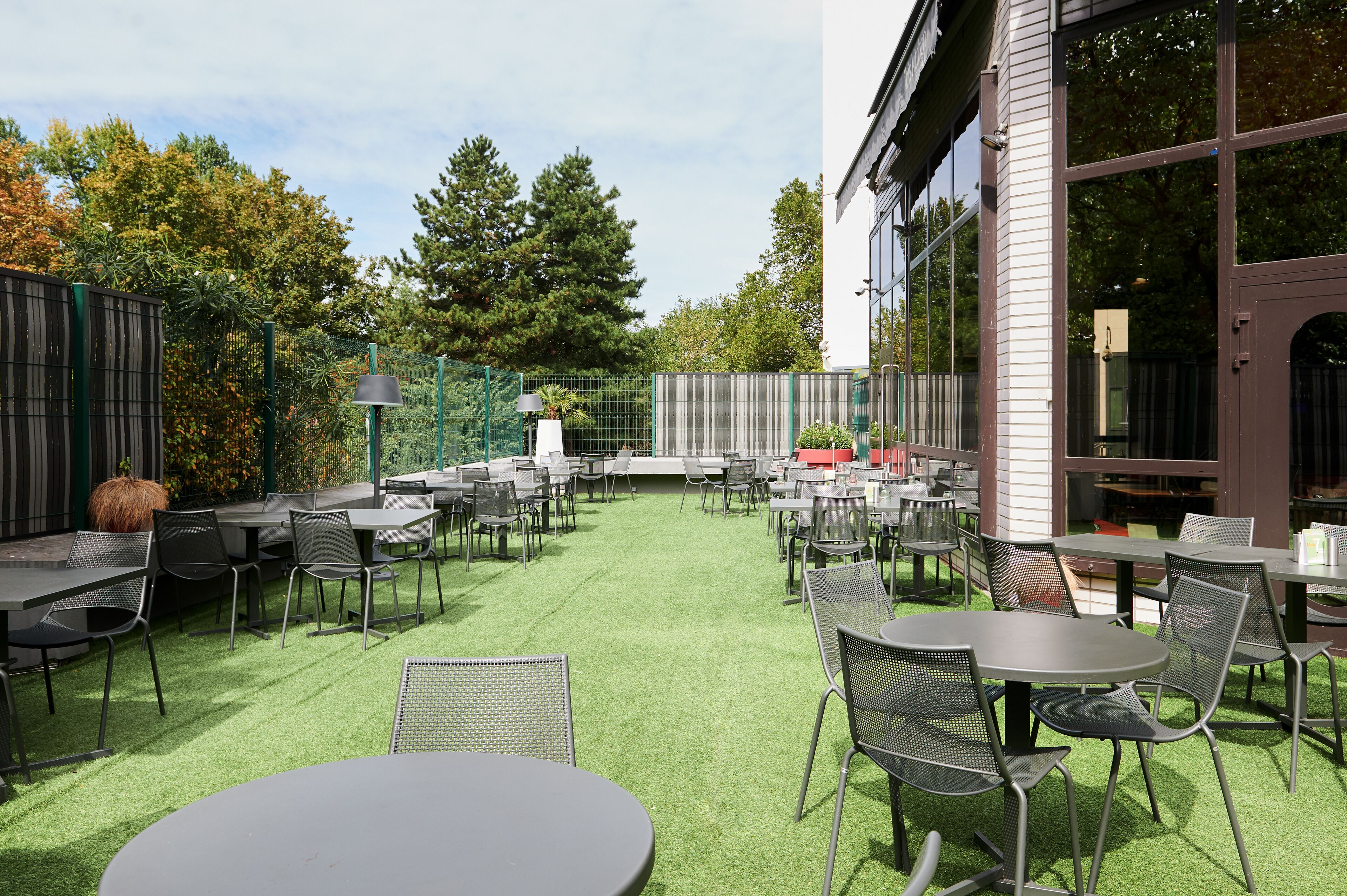 Un patio al aire libre con mesas y sillas, rodeado por una cerca verde y árboles.