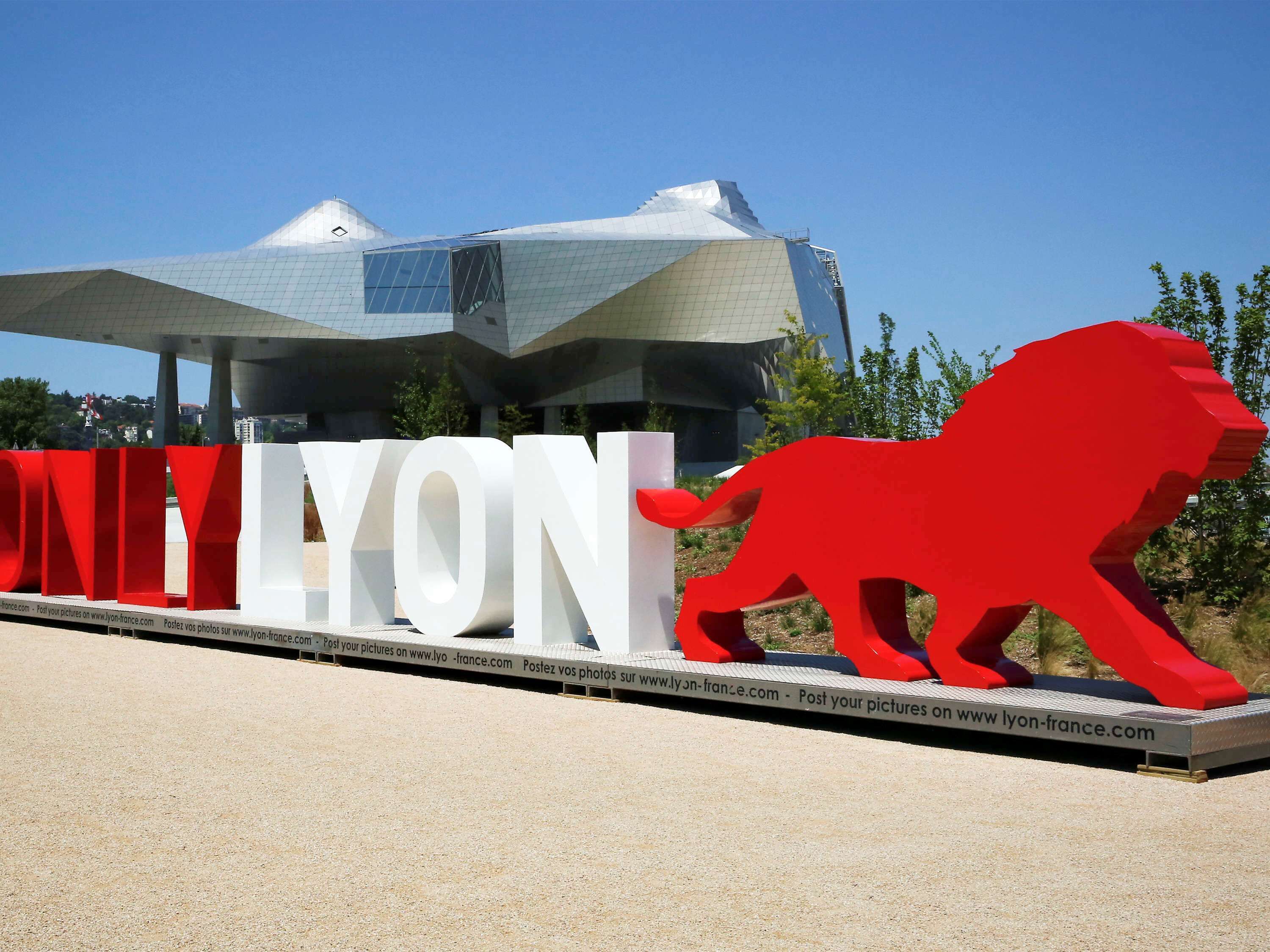 Un cartel grande con la inscripción 'LYON' y una escultura de león rojo frente a un edificio moderno.