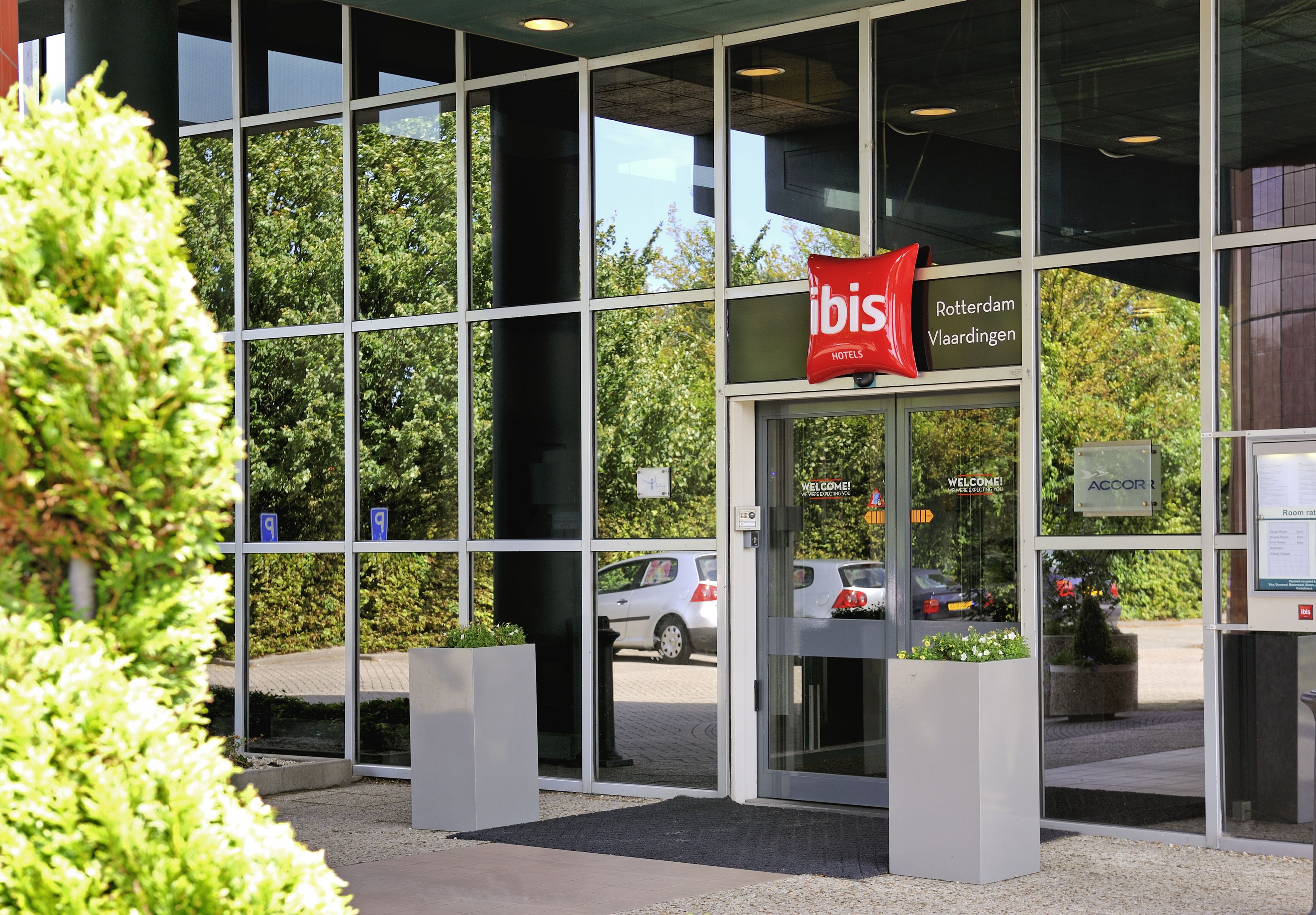 Una entrada de hotel moderna con fachada de vidrio, un letrero rojo de 'ibis' y un cartel que dice 'Bienvenido'.