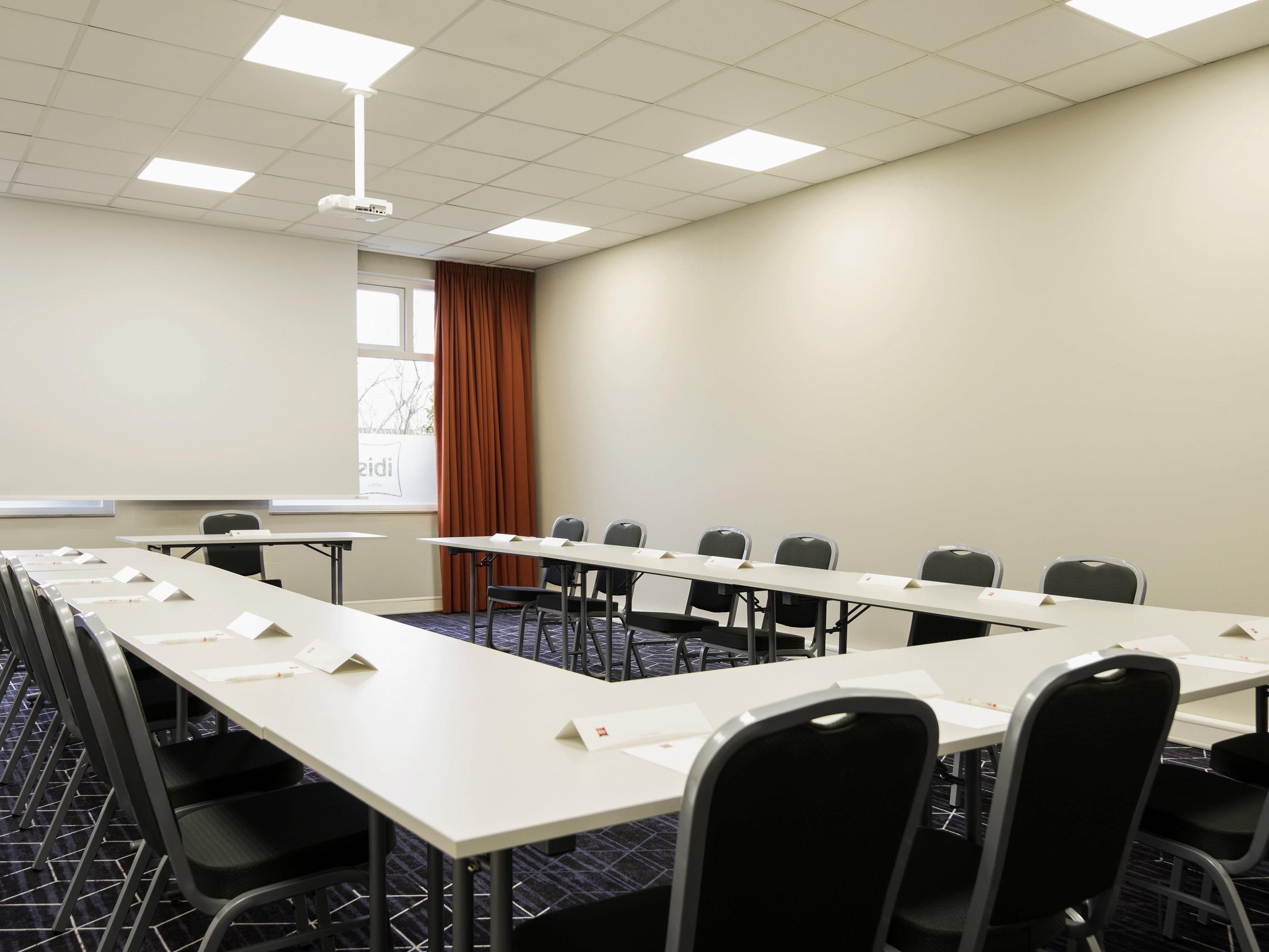A conference room with a long table, chairs, a projector screen, and a window with curtains.