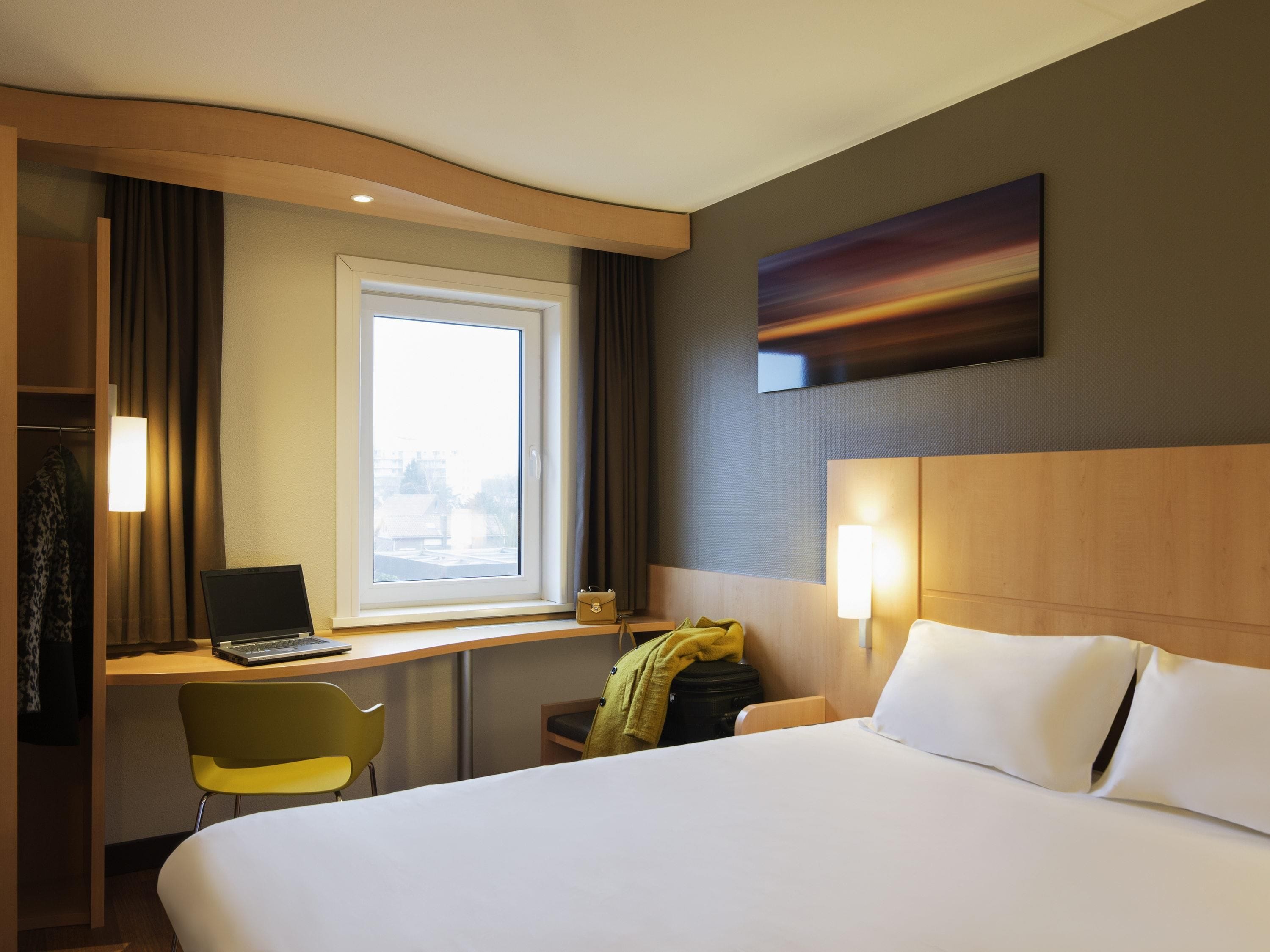 A hotel room with a bed, a desk with a laptop, a chair, a window, and a painting on the wall.