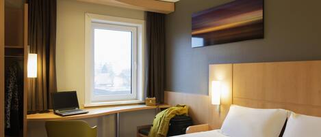 A hotel room with a bed, a desk with a laptop, a chair, a window, and a painting on the wall.