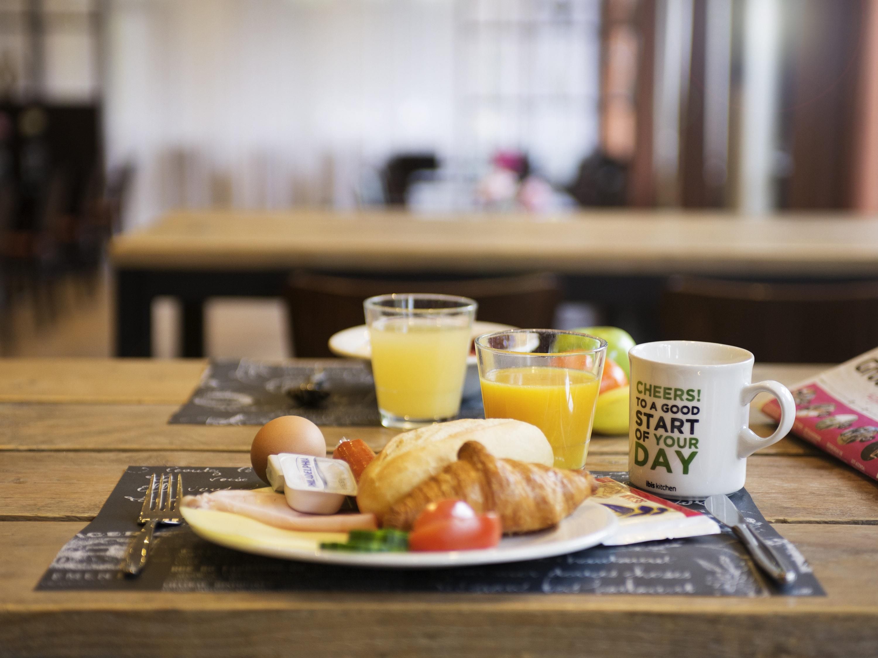 En morgenmadsopsætning med en croissant, et glas appelsinjuice, en kop med en opmuntrende tekst og en avis på et træbord.