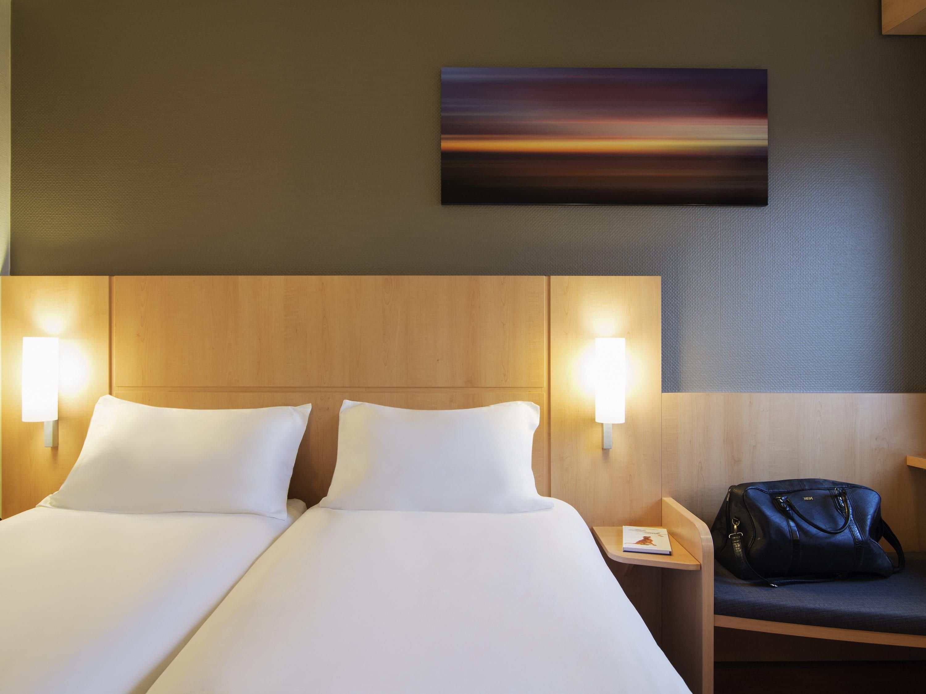 Ein Hotelzimmer mit Doppelbett, zwei Nachttischlampen, einem Nachttisch, einem Wandbild und einer schwarzen Handtasche auf einem Stuhl.