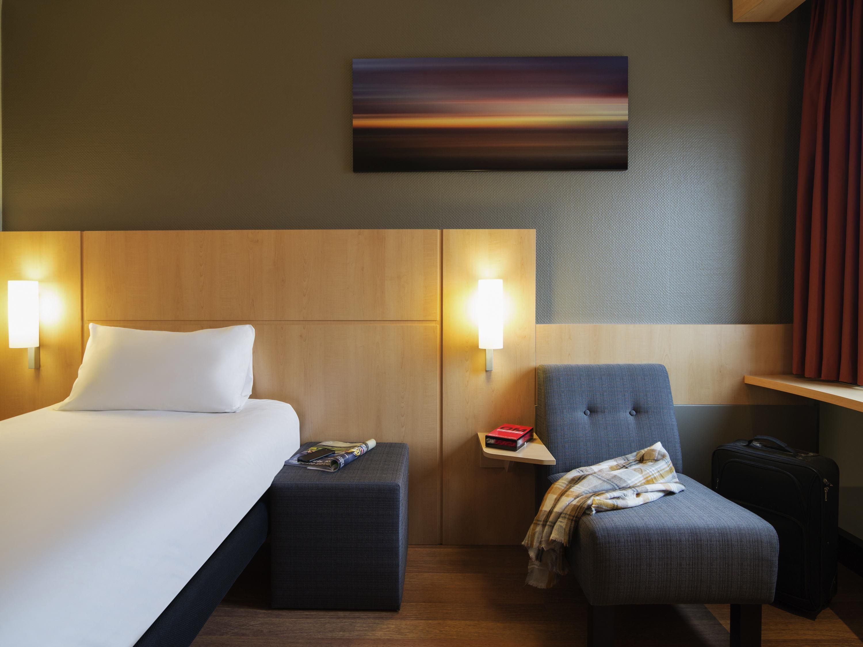 Una habitación de hotel con una cama, una silla, una mesita y una obra de arte colgada en la pared.
