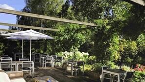 Un patio al aire libre con mesas y sillas, rodeado de follaje y una pérgola de madera.