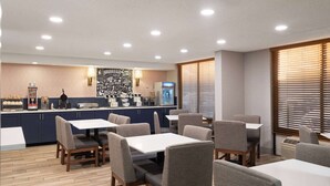 A modern hotel dining area with multiple tables and chairs, a kitchen in the background, and a wall with motivational quotes.