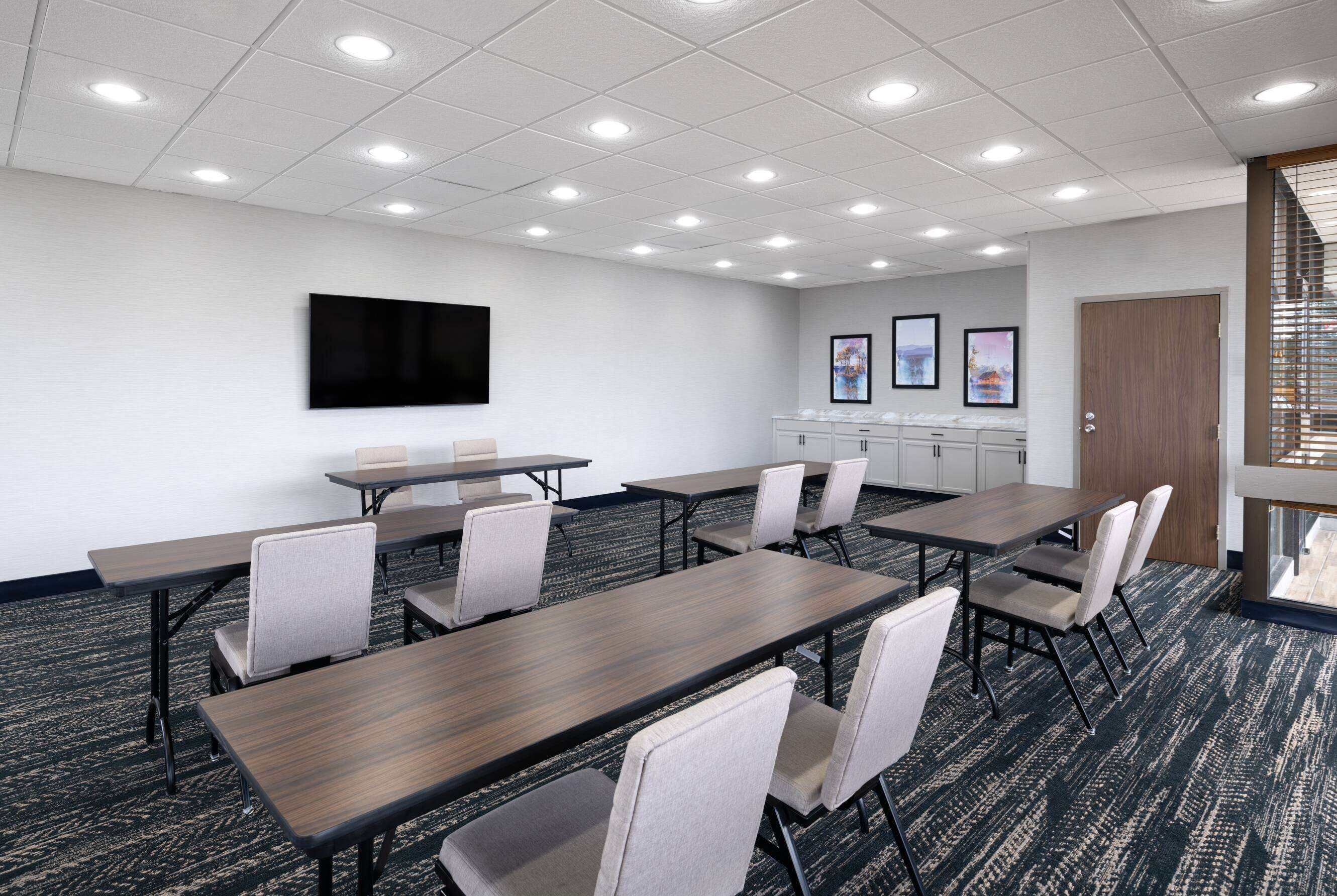 A conference room with multiple tables and chairs, a flat-screen TV, framed artwork on the wall, and a door with blinds.
