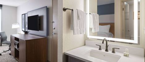 Baño de hotel con un espejo grande, un lavabo, tocador, televisor y una silla.
