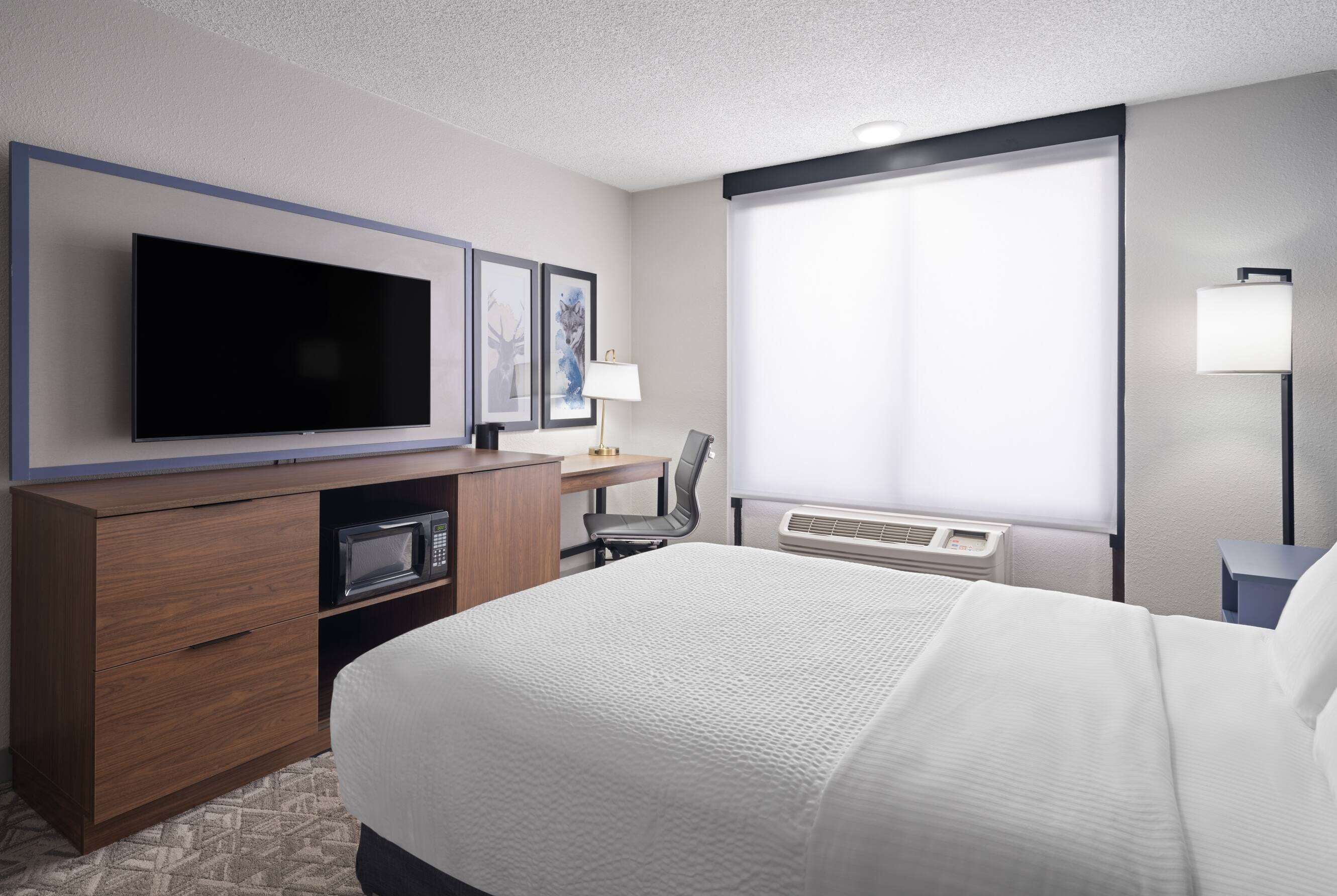 A hotel room with a large bed, a flat-screen TV, a desk with a chair, a microwave, a lamp, and a window with blinds.
