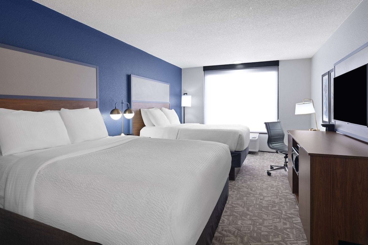 A hotel room with two beds, a desk, a chair, a TV, and a window.