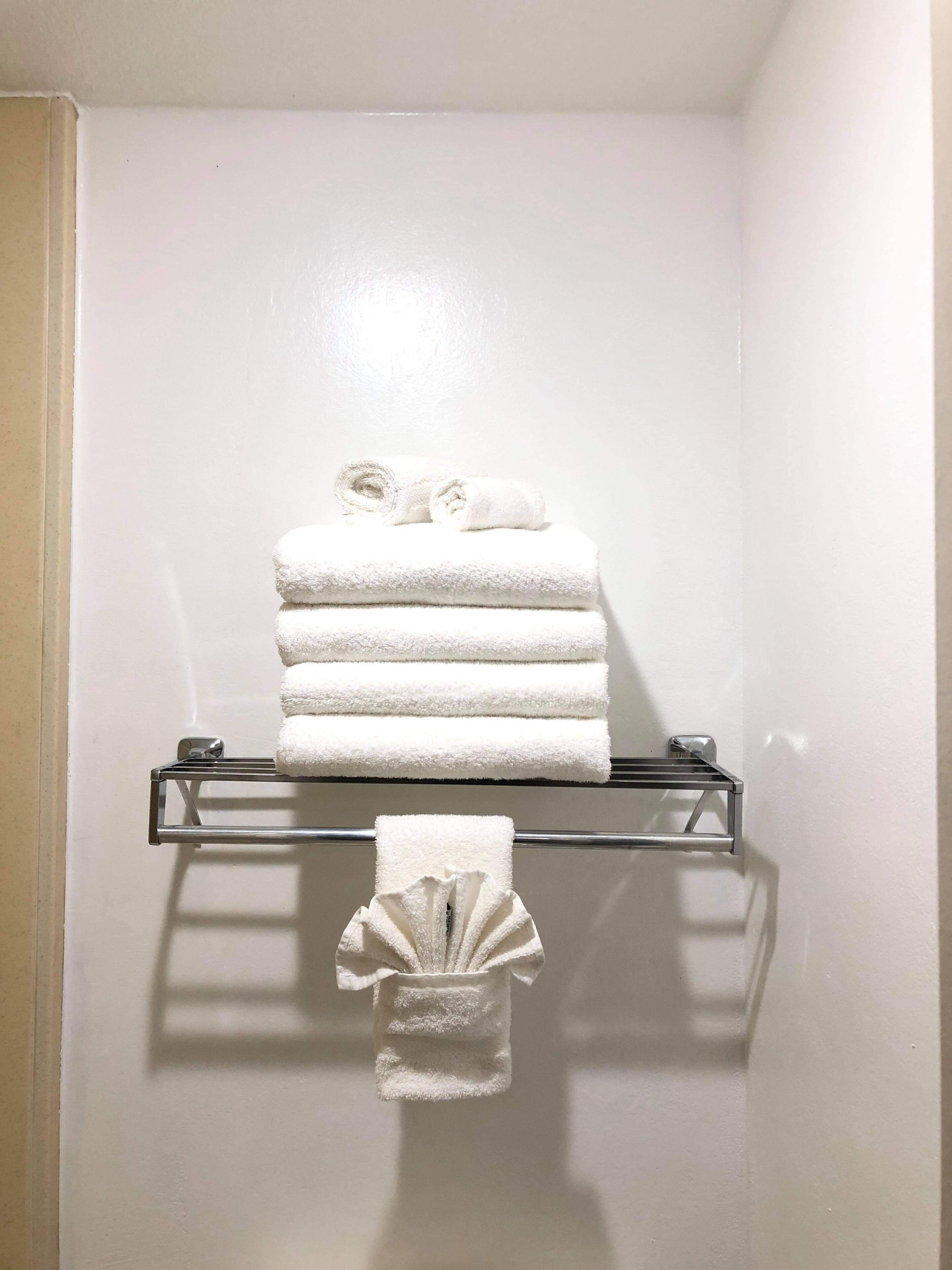 A bathroom with a white wall, a glass towel rack holding three folded towels, and a single towel hanging below the rack.