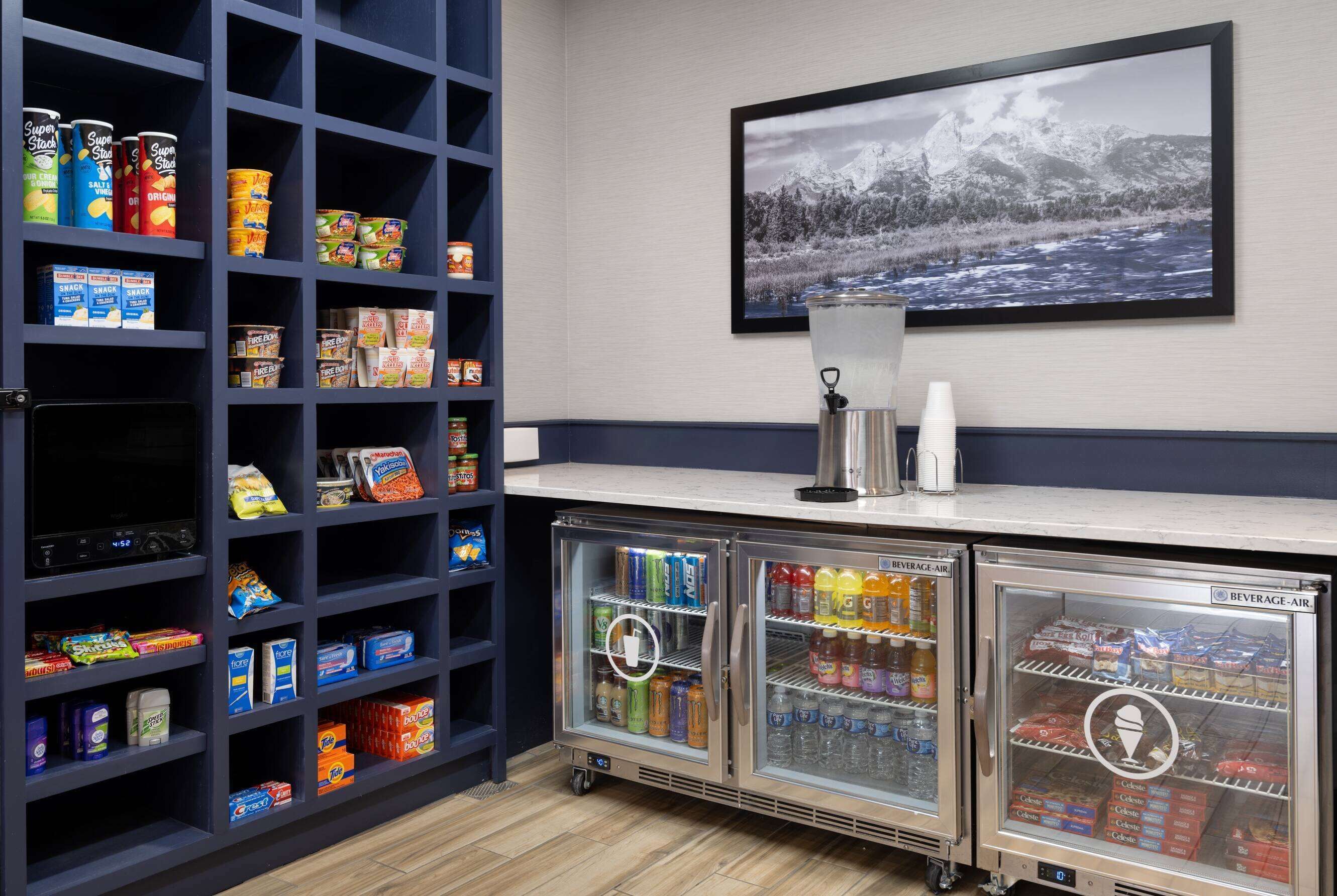 A kitchenette with shelves stocked with snacks and beverages, a refrigerator with drinks, and a framed picture of a snowy landscape on the wall.