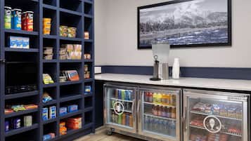 A kitchenette with shelves stocked with snacks and beverages, a refrigerator with drinks, and a framed picture of a snowy landscape on the wall.